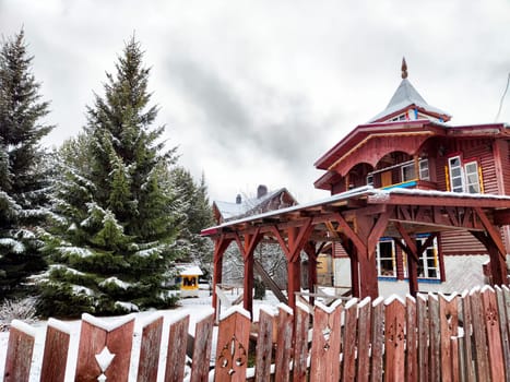 Charming wooden structures are blanketed in snow, surrounded by tall evergreen trees under a gray, overcast sky during winter.