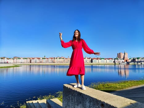 A woman smiles joyfully while standing on a concrete ledge near a river, showcasing vibrant architecture and clear blue sky.
