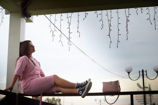 A woman in a pink outfit sits on gazebo railing, her legs dangling and her purse resting on the rail beside her