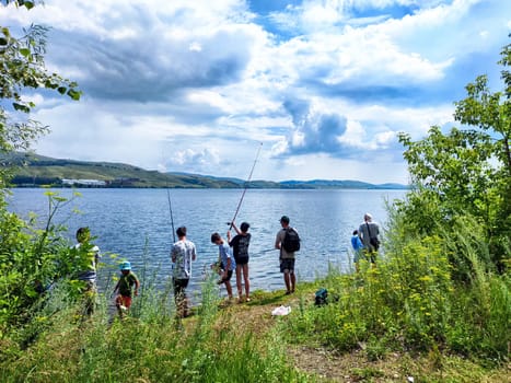 Abzakovo, Russia - July 18, 2024: Groups of individuals spend a sunny afternoon fishing by the lakeshore with scenic views and lush vegetation nearby.