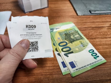 Kirov, Russia - October 08, 2024: A person holding a voucher for currency exchange next to a stack of 100 euro banknotes at a bustling exchange counter.