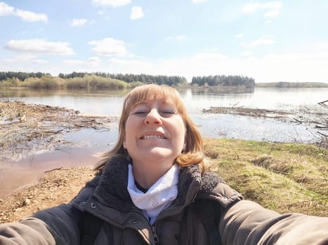 A woman in her 40s or 50s takes selfie outdoors near water on a sunny day. She is wearing a blue jacket and smiling brightly at the camera