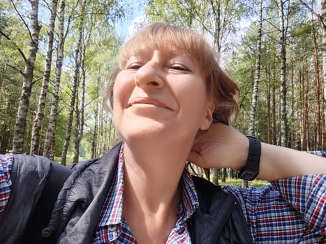 A middle-aged woman with blonde hair smiles while taking a selfie in a lush green forest surrounded by trees.