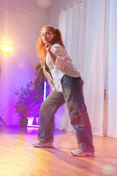 A young woman with curly red hair, wearing a white shirt and blue jeans, dances in a studio. The background is blurred, featuring a brick wall, white curtains, and colored lights.