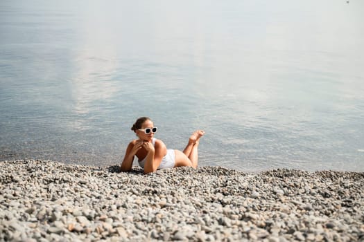 A woman is laying on the beach wearing sunglasses and a white tank top. She is looking at the camera