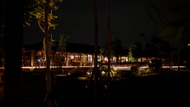 Nighttime view of a lit-up building with warm lights, surrounded by darkness and silhouetted foliage.