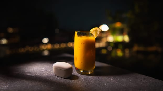 Glass of orange juice with a slice of lemon on a table at night, with blurred lights in the background.