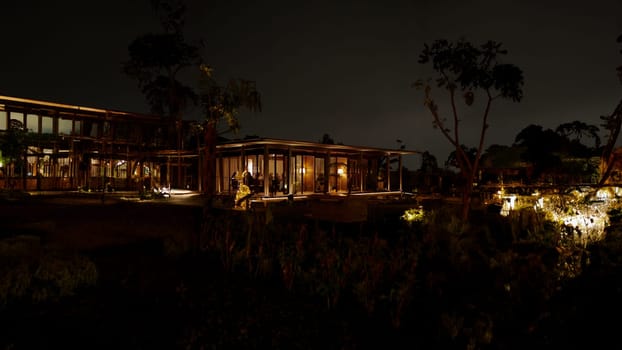 Nighttime view of a modern house with illuminated interiors and exterior lights, surrounded by dark silhouettes of trees in the dark night