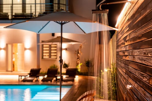 Outdoor shower with a sleek wooden wall next to a swimming pool illuminated at night. Concept of modern backyard luxury combining comfort and elegance