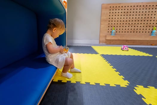 Small child sits on a cozy bench in a colorful playroom, holding a toy and exploring the environment. Concept of joyful play, early education, and creative spaces for kids
