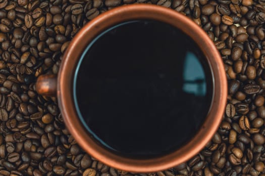 Overhead view of a clay cup filled with black coffee surrounded by roasted coffee beans, creating a rustic and textured composition, no people.
