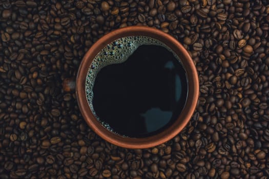 Overhead close-up view of a brown ceramic cup filled with black coffee placed on a bed of roasted coffee beans.