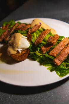Gourmet steak salad with poached eggs and crispy bread on a white plate, dark moody lighting.