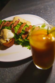 a glass of orange juice next to a plate of chicken meat and green salad on the table in the cafe at night