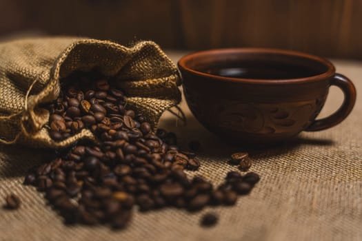 A brown ceramic cup of black coffee placed next to a burlap sack spilling roasted coffee beans on a textured burlap surface.