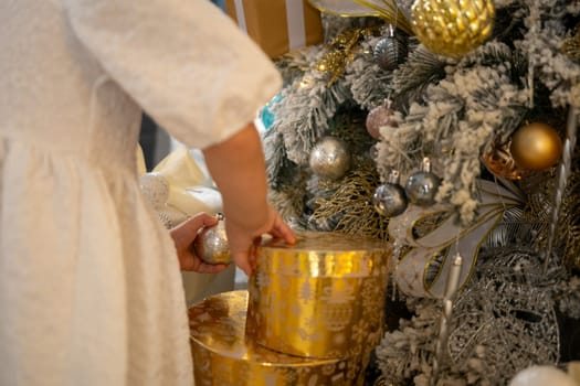 A woman is opening a gold box with a silver ball inside. The box is decorated with gold and silver ornaments, and the woman is wearing a white dress. The scene is set in a room with a Christmas tree