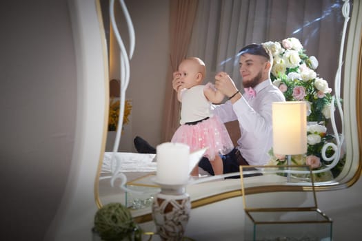 A father embraces his baby daughter near a floral arrangement in a cozy indoor setting.