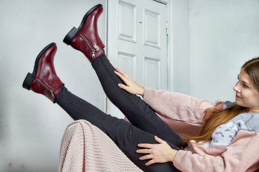 Stylish model effortlessly poses with legs extended, highlighting fashionable burgundy ankle boots in a chic room.