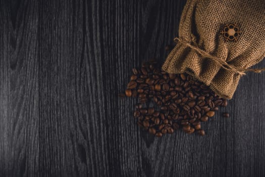 A burlap sack spilling roasted coffee beans onto a textured dark wooden surface with a rustic aesthetic.