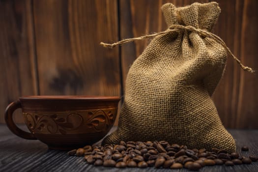 Burlap sack filled with coffee beans, a decorative clay cup, and spilled beans on a textured wooden table with a warm background, rustic style, no people.