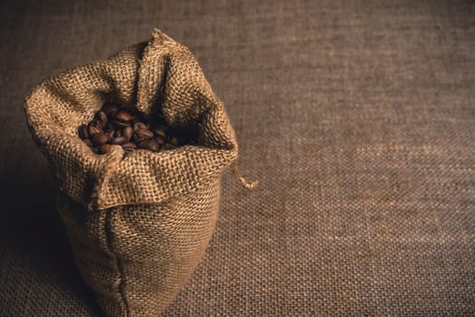 A rustic burlap sack filled with roasted coffee beans placed on a textured burlap fabric background.