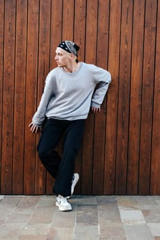 A young man in a grey sweatshirt and black pants with a black bandana on his head is leaning against a brown wooden wall.