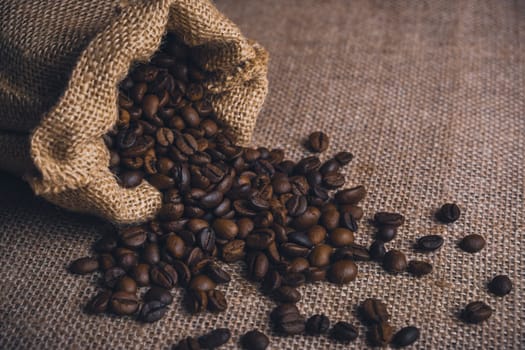Burlap sack tipped over with roasted coffee beans spilling onto a textured burlap fabric background.