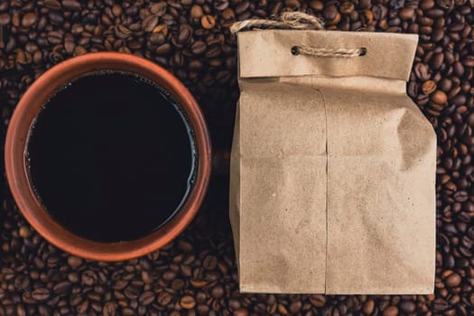 A top view of a ceramic coffee cup filled with black coffee next to a kraft paper bag, surrounded by roasted coffee beans.