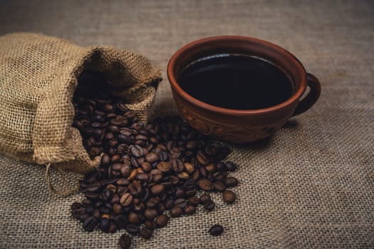 A rustic brown ceramic cup filled with black coffee next to a burlap sack spilling roasted coffee beans onto a burlap fabric surface.