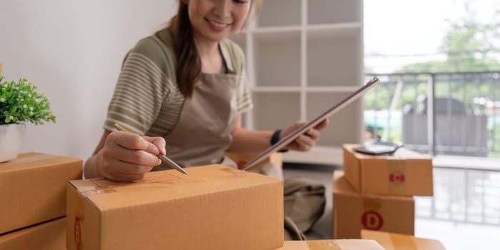 Woman asian is writing a list of customer on paper before shipping to them, she runs an ecommerce business on websites and social media. Concept of selling products online.