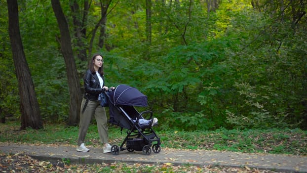 A young mother walks through the forest with a sleeping child in a stroller. A woman with glasses walks in an autumn park. 4k