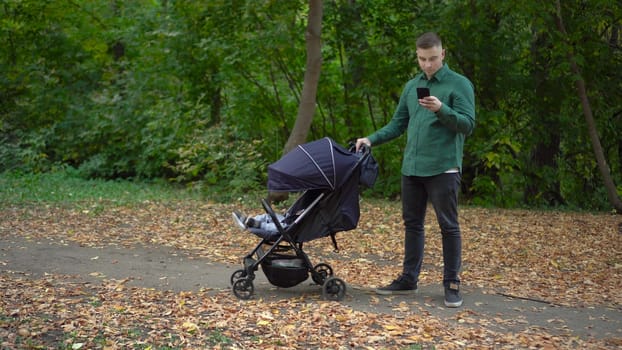 A young father is texting on a smartphone and rocking his child in a stroller. A man with a phone in the autumn forest. 4k