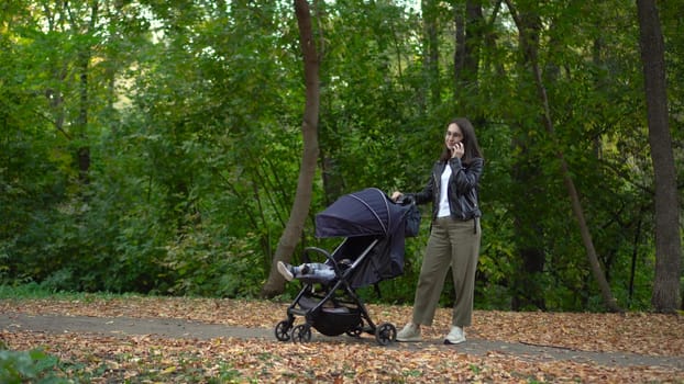 A young mother is talking on the phone and rocking her baby in a stroller. A woman with glasses speaks on the phone in the autumn forest. 4k