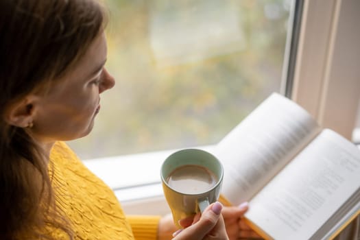 Young beautiful woman sitting by the window yellow knitted sweater read book, daily planner, notepad. Relax concept. Hold cappuccino glass of coffee with white foam. Text is out of focus