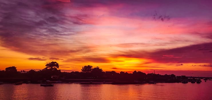 The photo captures the beauty of a sunset over an Ocean. The sky is filled with orange, pink, and purple clouds, and the Ocean in Nusa Dua, Bali reflects the colors of the sky. Boats are on the river, and trees. High-Quality Photo.