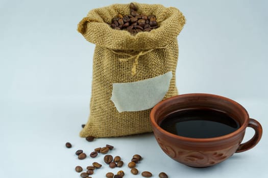 Burlap sack filled with roasted coffee beans, featuring a blank label, next to a decorative coffee cup with black coffee on a white background.