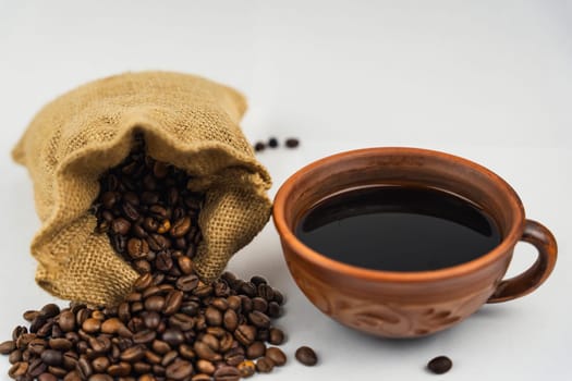 Coffee beans spilling from burlap sack next to a clay cup filled with black coffee on white background, rustic and natural style.