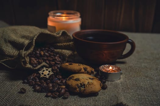 A rustic brown ceramic cup of black coffee paired with chocolate chip cookies, burlap sack spilling coffee beans, and lit candles on a burlap fabric background.