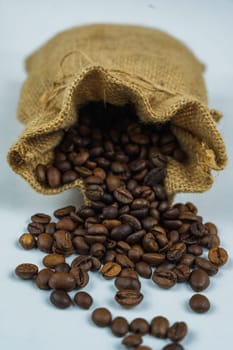 Rustic burlap sack with roasted coffee beans spilling out onto a clean white background, highlighting the texture and natural material.