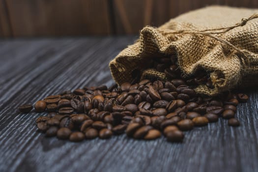 Roasted coffee beans spilling from a rustic burlap sack onto a textured wooden table with a warm, dark background, no people.