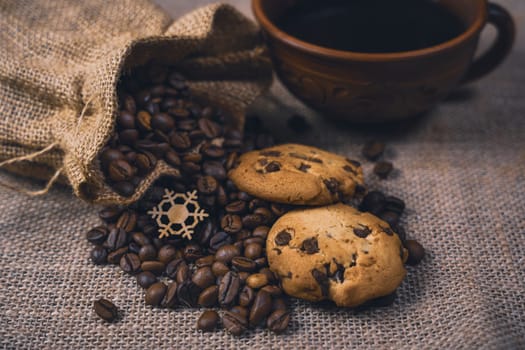 Rustic still life featuring a burlap sack spilling coffee beans, chocolate chip cookies, and a ceramic cup of coffee on a burlap background.