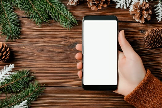A hand holding smartphone with blank screen surrounded by pinecones and greenery creates cozy winter atmosphere