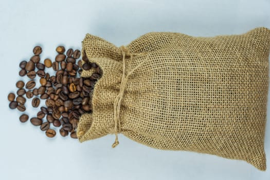 Burlap sack with roasted coffee beans spilling onto a white surface, high-angle view, isolated, no people, showcasing natural packaging.