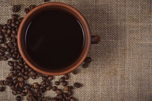 Rustic composition featuring an overhead view of a ceramic coffee cup with black coffee and scattered coffee beans on a burlap fabric background.