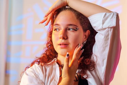 A young woman with red, curly hair is posing against a light background. She is wearing a white button-up shirt with a black choker and has on decorative, long nails.