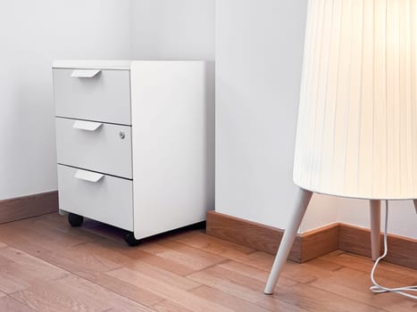 Minimalist white cabinet and modern lamp in bright corner of wooden floored room.