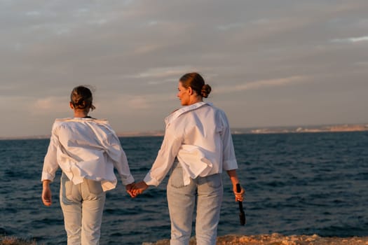 Two women, dressed in white shirts and blue jeans, walk hand-in-hand along a rocky shore, gazing at the ocean at sunset.