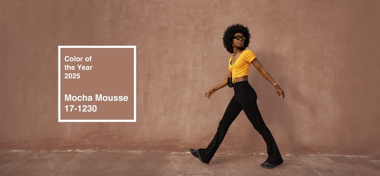 A model confidently walks against a textured backdrop wearing a vibrant yellow top and black pants. The warm light highlights the mocha mousse color trend for 2025, creating a fashionable vibe.