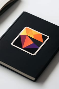 A square sticker featuring a vibrant geometric design rests on a matte black notebook. photographed in a professional studio, this setup highlights the contrast between the colorful sticker and the sleek notebook, perfect for showcasing print designs.