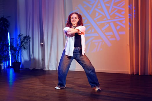 A young woman with curly brown hair is dancing in a studio. She is wearing a white shirt, blue jeans, and white sneakers.